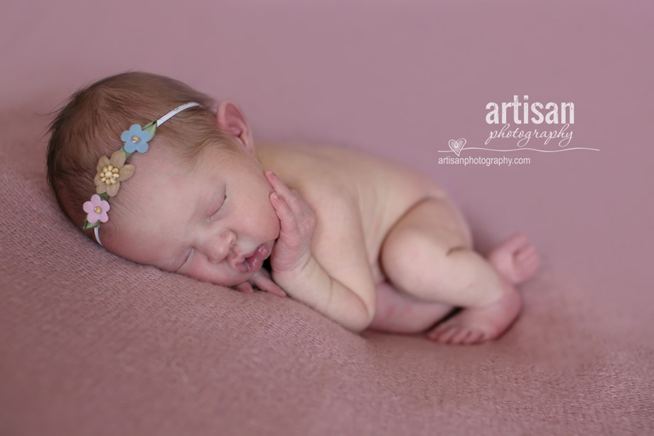 baby girl laying on blanket with flower headband in dusty pink background at the artisan studio