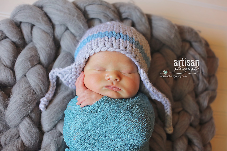 newborn baby boy laying on a chunky grey blanket with a grey and blue aviator hat in Carlsbad california