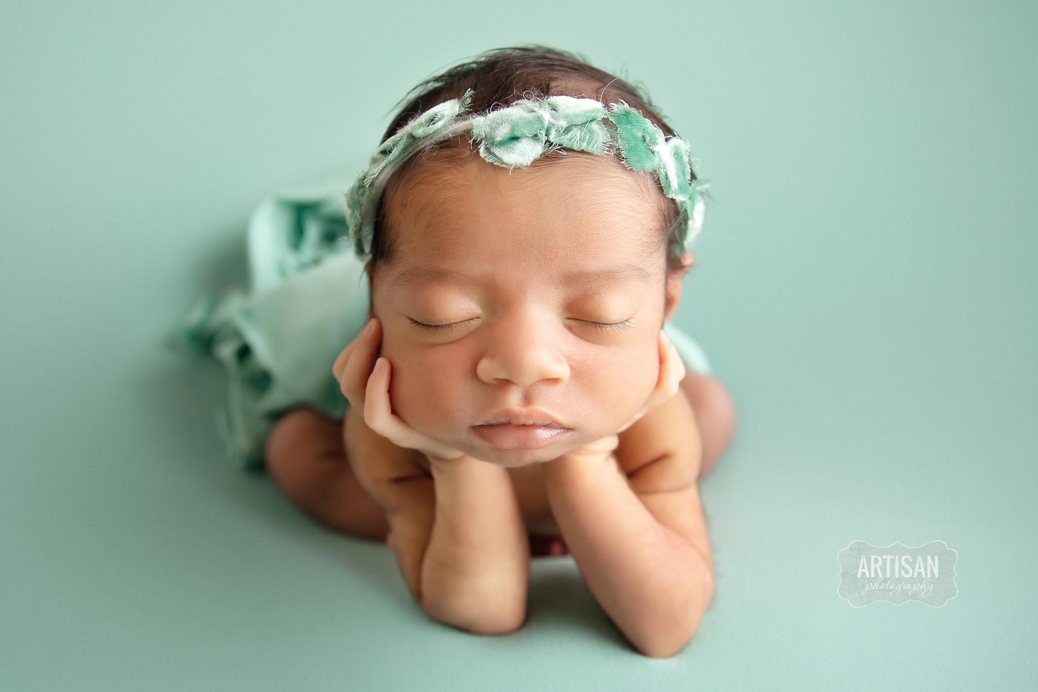 Newborn baby girl posing with hands on chin with a mint green backdrop.