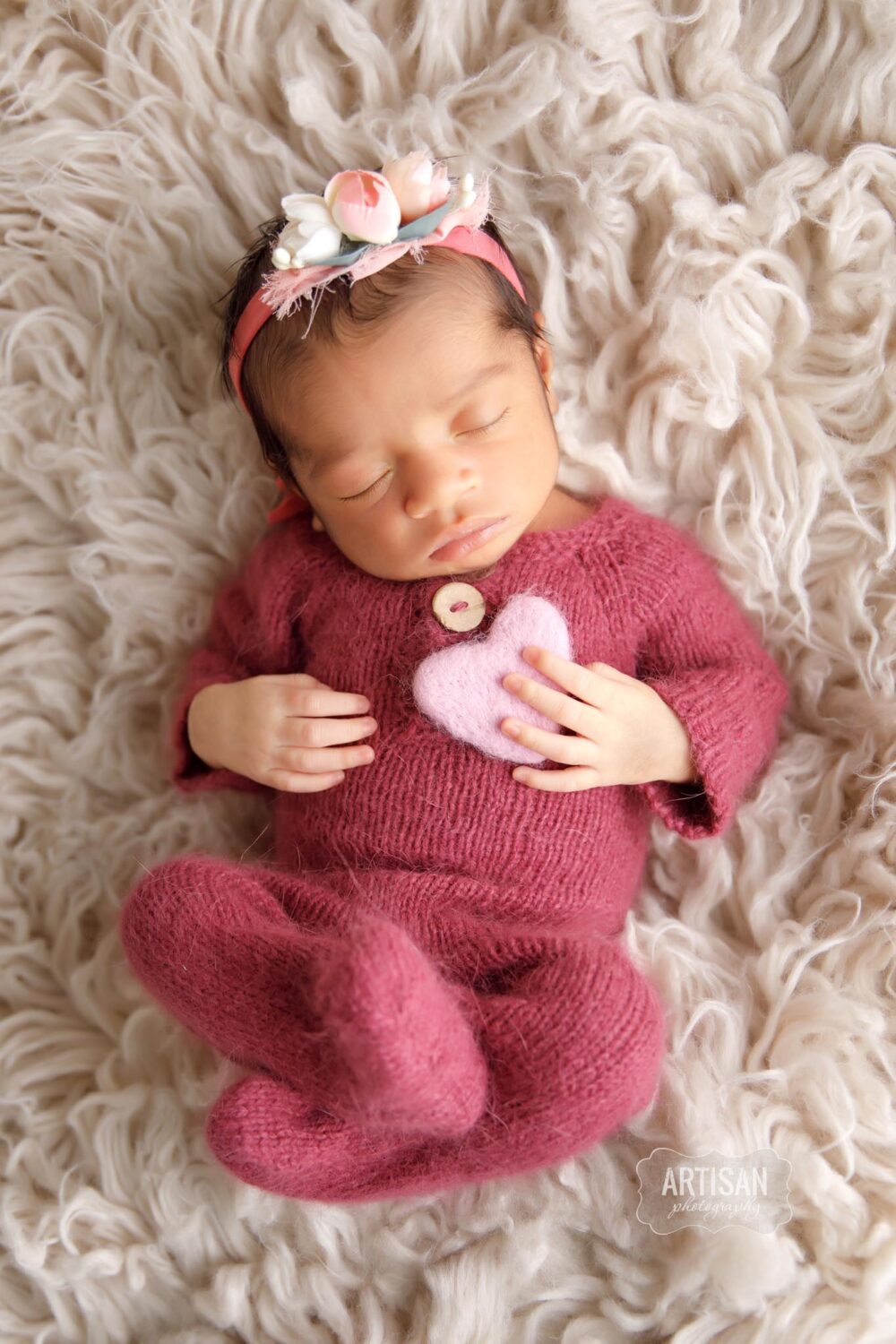 Newborn baby girl wearing a pink outfit holding a light pink lavender felted heart on cream flokati fur 